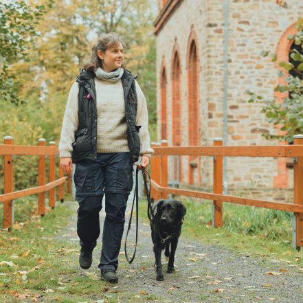 Eine Frau führt einen Hund an der Leine über einen naturbelassenen Weg entlang eines Holzgeländers. Im Hintergrund steht ein Gebäude aus San
