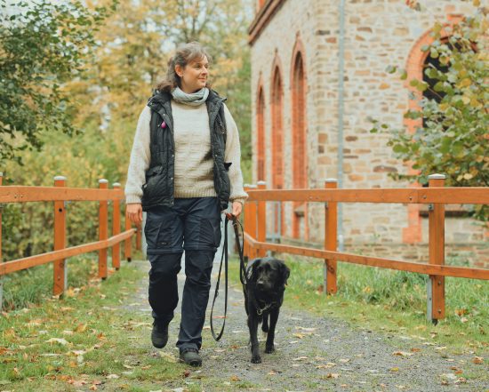 Eine Frau führt einen Hund an der Leine über einen naturbelassenen Weg entlang eines Holzgeländers. Die Szene wirkt ruhig und entspannt.