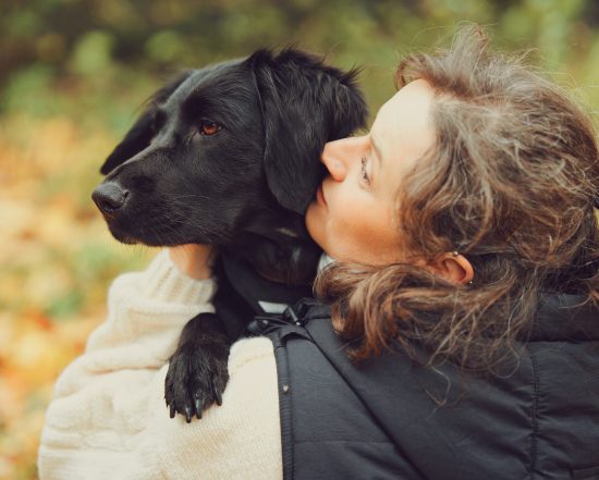 Frau umarmt und kuschelt einen schwarzen Hund im herbstlichen Umfeld.